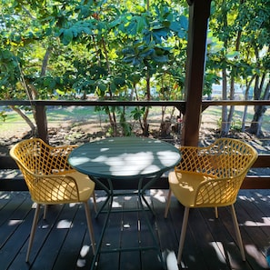 Una terraza de madera con dos sillas amarillas y una mesa, rodeada de follaje y árboles.