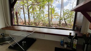 A kitchen with a window overlooking a beach, a sink, a dish rack, and kitchen appliances. - Beach bungalow: Barracuda (Le Carbet)