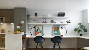 A modern kitchen with a white countertop, two black chairs, and a wooden stool. There are two framed artworks, a clock, and a small plant on the shelf. - For Students Only Studios in Plymouth (Plymouth)
