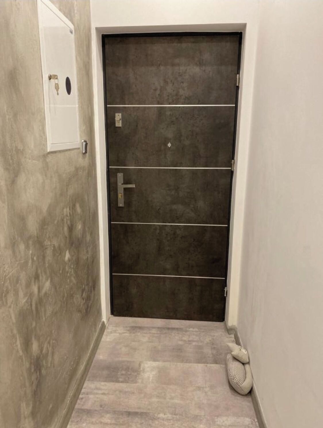 Ein Korridor mit einer dunkelholzigen Tür, einer weißen Wand und einem strukturierten Boden.