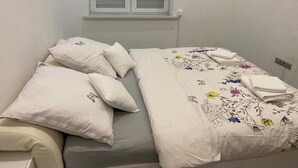 Una cama individual con una colcha estampada y dos almohadas, una ventana con contraventanas y un interruptor de luz en la pared.