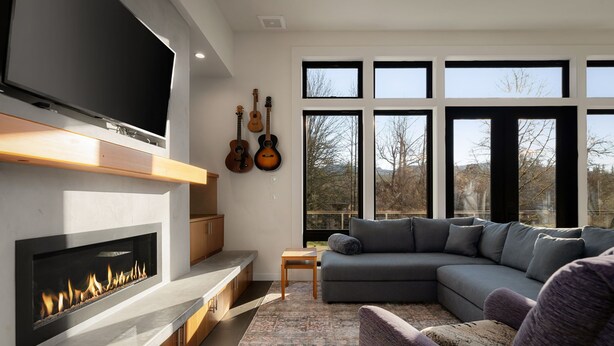 A modern living room with a fireplace, a large flat-screen TV, a grey sectional sofa, and a view of the outdoors through large windows. - Bellingham Adventure Home: 4BR Retreat w/ Yard Near Trails (Bellingham)