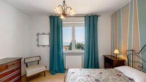 Une chambre à coucher comprenant un lit, une commode, une chaise, une fenêtre avec des rideaux, un radiateur et une vue sur l’extérieur.