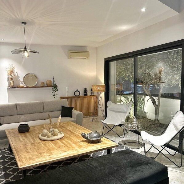 Ein modernes Wohnzimmer mit einer Couch, einem Holztisch und einem Glastisch mit Stühlen. Ein großes Fenster bietet Blick auf Bäume und ein Gebäude.