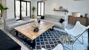 A modern living room with a large sofa, a coffee table, and a white armchair. There are potted plants and a hanging light fixture. - La Casa Verde , Duplex in the Heart of La Marsa (La Marsa)