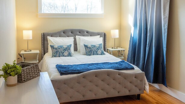 A bedroom with a tufted headboard, two bedside tables with lamps, a window with a view of bare trees, and a blue curtain. - Ann Inn - Downtown New Milford Apartment (New Milford)