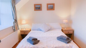 A bedroom with a bed, two nightstands, two lamps, two framed pictures, and a window with curtains. - Tuskar Holiday Cottage, Mill Road Farm (Kilmore Quay)