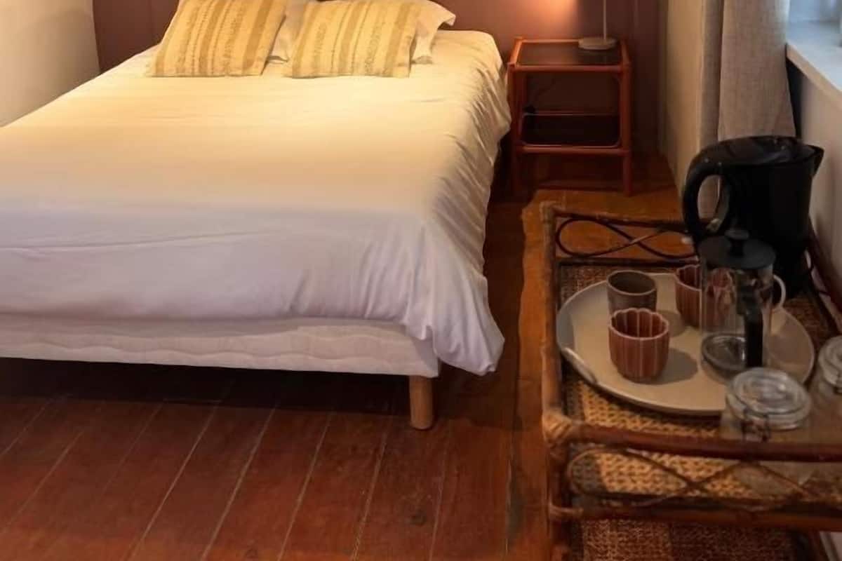 Ein ordentlich bezogenes Bett mit Kissen, ein Nachttisch mit Kaffeemaschine und Tassen sowie zwei gerahmte Bilder an der Wand.