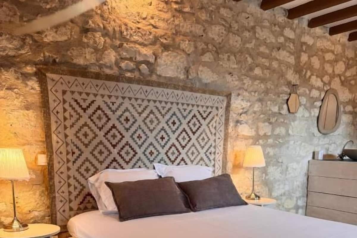 Ein Schlafzimmer mit Steinwand, einem gemusterten Kopfteil, einem Bett mit weißen Leinen, zwei Nachttischen mit Lampen und einer Holzkommode.