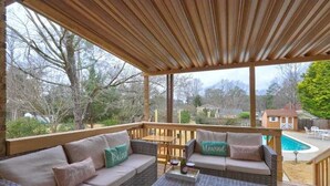 A covered outdoor seating area with wicker furniture and cushions, overlooking a backyard with a swimming pool and trees. - Luxury 3BR Getaway 2 Master Suites Pool & More, Great Location (Stone Mountain)