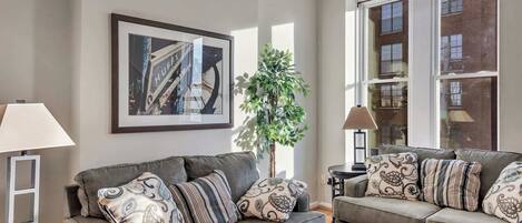 A living room with two grey sofas, patterned cushions, a framed abstract painting, a potted plant, and a large window with a view of a building.