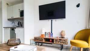 Ein modernes Wohnzimmer mit einem an der Wand befestigten Flachbildfernseher, einem Holztisch, einem senffarbenen Sessel und einer Küchenzeile mit weißen Schränken und einer Mikrowelle.