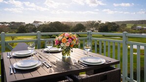 Ein Tisch für zwei Personen, gedeckt mit Blumen, Tellern und Weingläsern, auf einer Holzterrasse mit Blick auf eine grüne Landschaft.