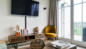 Ein Wohnzimmer mit einem großen Flachbildfernseher an der Wand, einem Holzkonsolentisch mit Dekorationen, einem senffarbenen Sessel und einer Glasschiebetür, die auf einen Balkon führt.