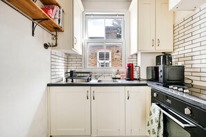 A compact kitchen with white cabinets, a black countertop, a window, and a brick wall. - The Vintage Cove (Greater London)