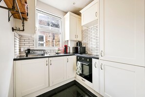 A kitchen with white cabinets, a black countertop, a window, and a brick wall. - The Vintage Cove (Greater London)