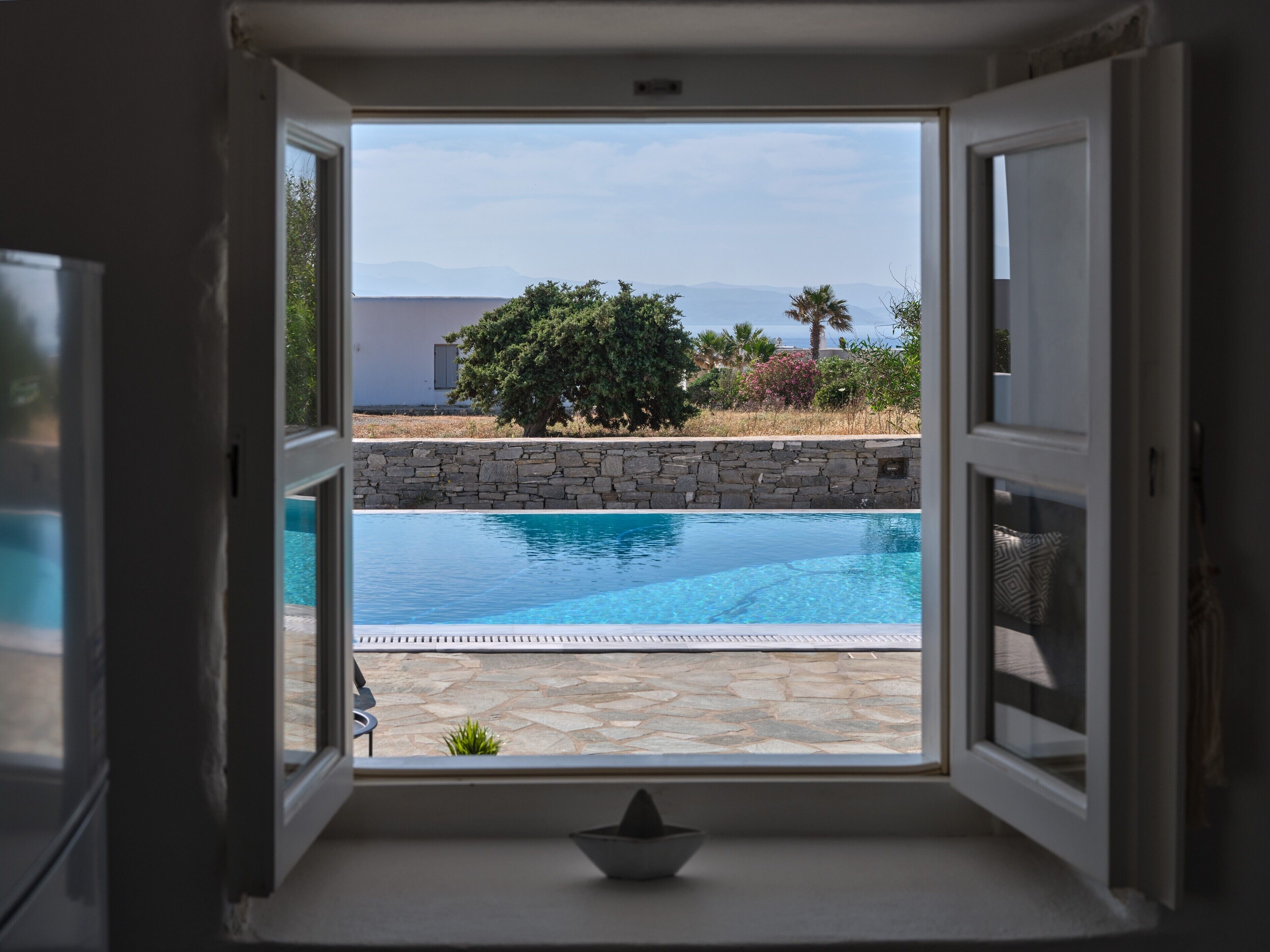 A view through an open door to a swimming pool with a stone wall and greenery in the background.