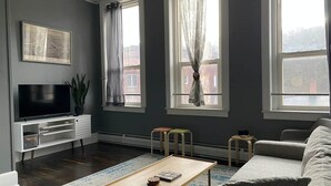 A modern living room with a flat-screen TV, a wooden coffee table, a grey sofa, and a patterned rug. - The Honesdale Loft - downtown historic Main St (Honesdale)