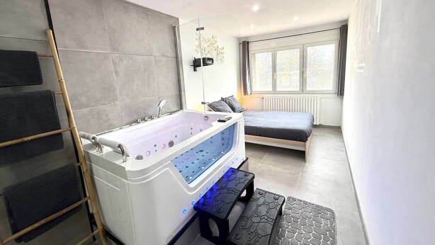 Une salle de bain moderne avec une grande baignoire, une douche et des toilettes. La pièce adjacente contient un lit et une fenêtre.