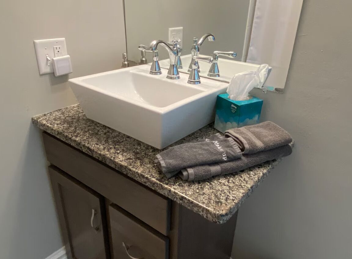 Une salle de bain avec un lavabo blanc, trois robinets, une boîte de mouchoirs et une serviette pliée posée sur le comptoir.