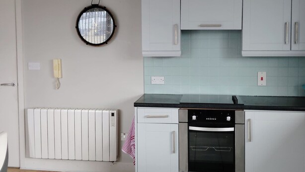A kitchen with white cabinets, a black countertop, a round wall clock, a white radiator, and a tiled floor. - 2 Bedroom Apartment 4-6 People-coastal Comfort (Balbriggan)