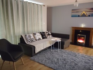 A living room with a sofa, a small round table, a chair, a fireplace, and a painting on the wall. - 2 Bedroom Apartment 4-6 People-coastal Comfort (Balbriggan)