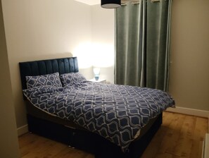 Una cama con cabecera oscura, una colcha estampada y una mesita de noche con una lámpara.