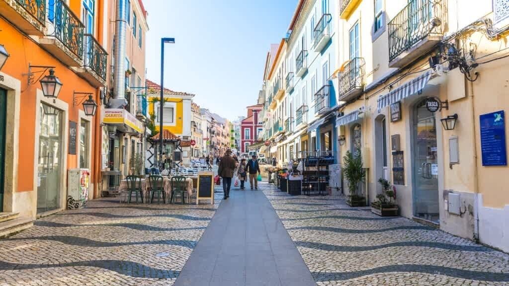 A cobblestone street lined with colorful buildings, some with outdoor seating and shops.