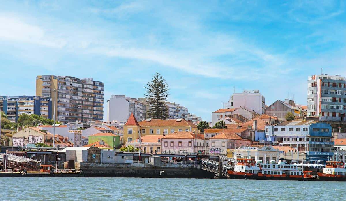 A waterfront cityscape with colorful buildings, a prominent tree, and a dock with boats.