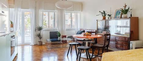 A well-lit room with a wooden floor, a round dining table with chairs, a sofa, a bed with a yellow blanket, a cabinet with plants, and a large window with curtains.