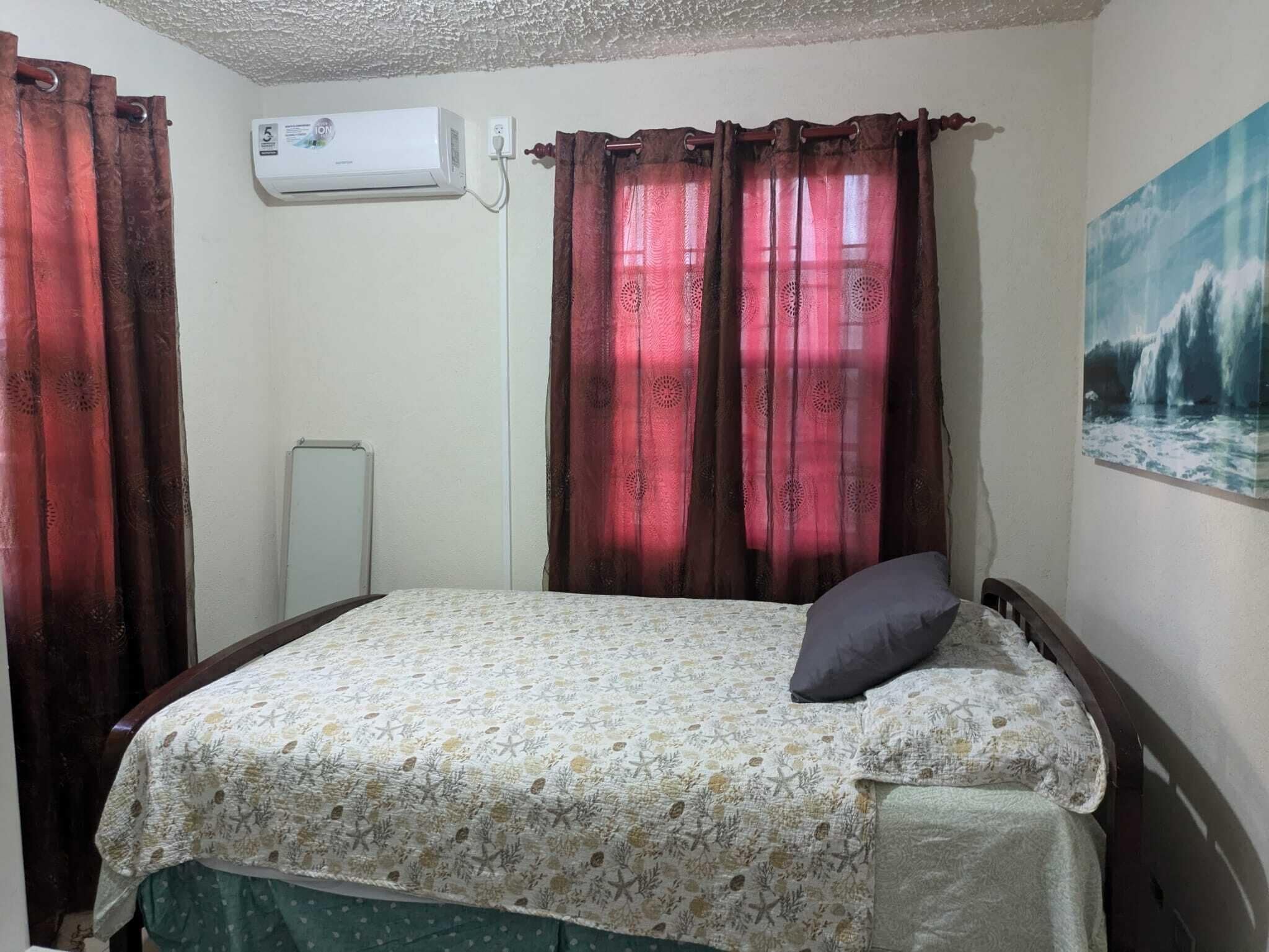 Un dormitorio con una cama, un cojín, un cuadro y cortinas.
