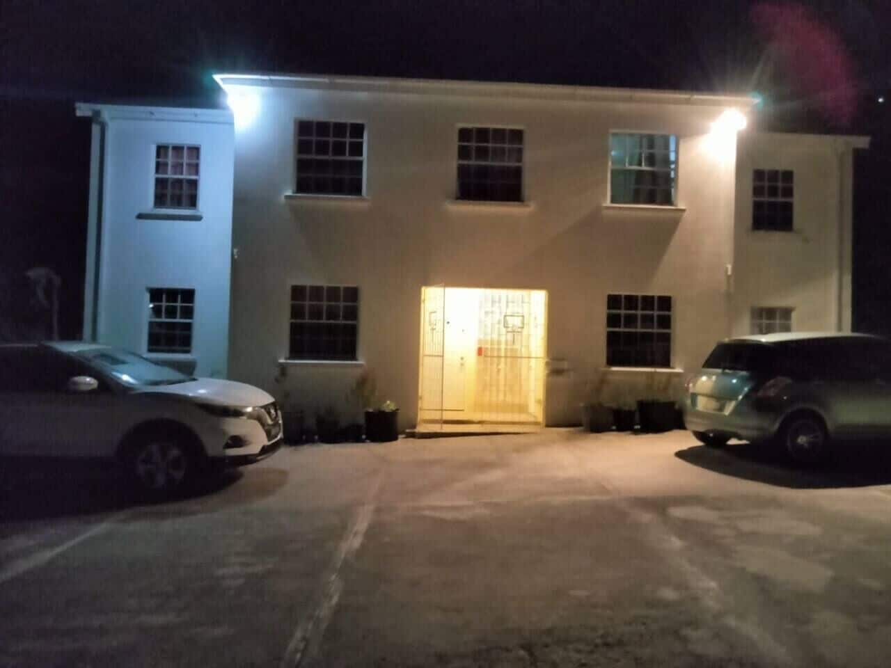Un edificio con entrada central, dos coches estacionados al frente y un pequeño jardín a la izquierda.