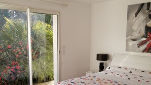 Quarto com cama, mesa de cabeceira, luminária, porta de vidro deslizante para um jardim e um quadro na parede.
