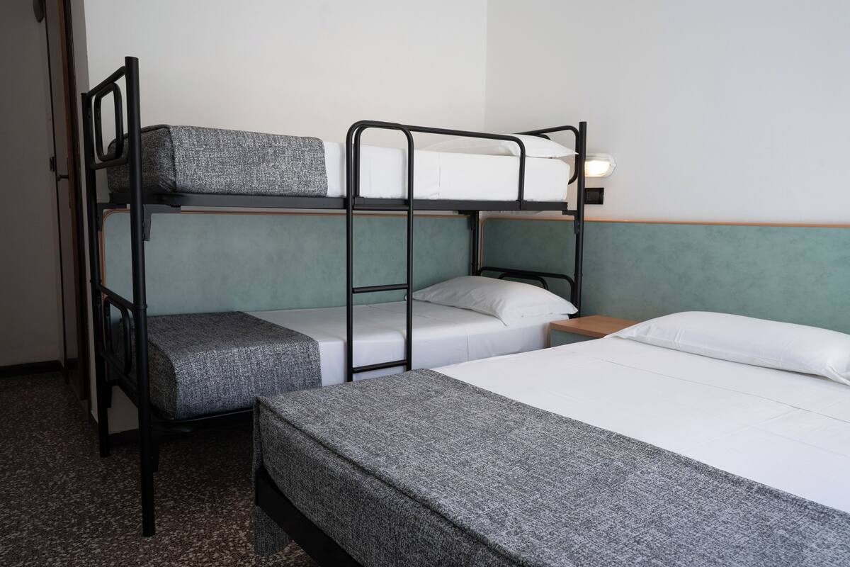 A bunk bed room with a green headboard, a single bed with a grey blanket, and a nightstand with a lamp.