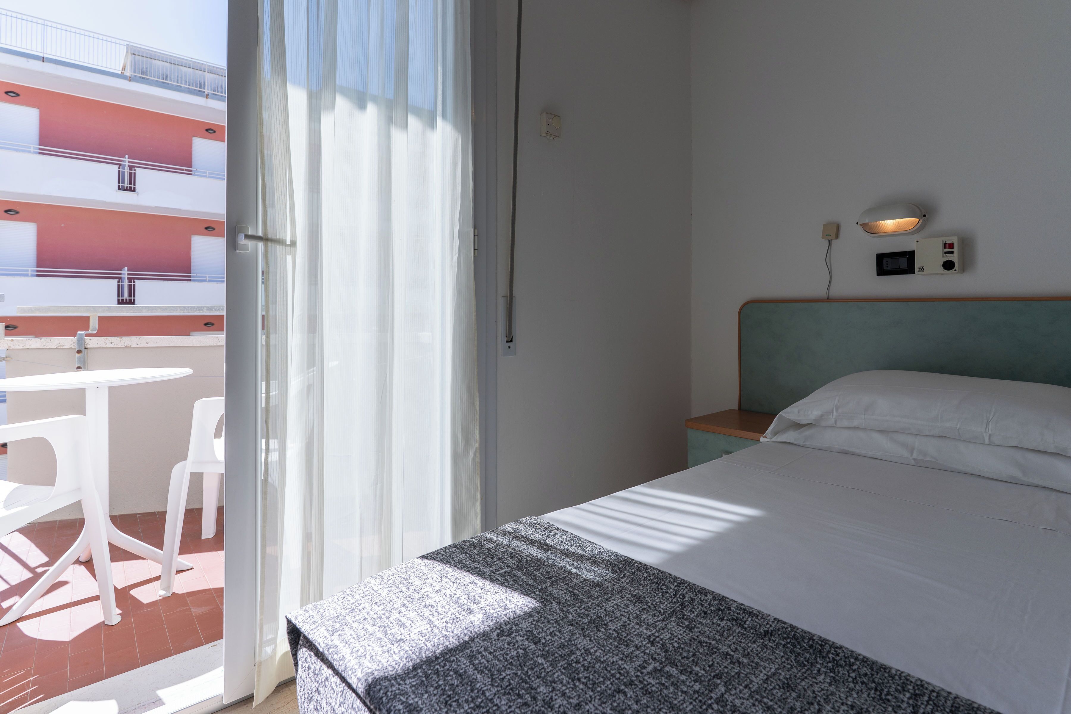 A hotel room with a bed, a bedside table, a wall-mounted lamp, and a view of an outdoor balcony with chairs.