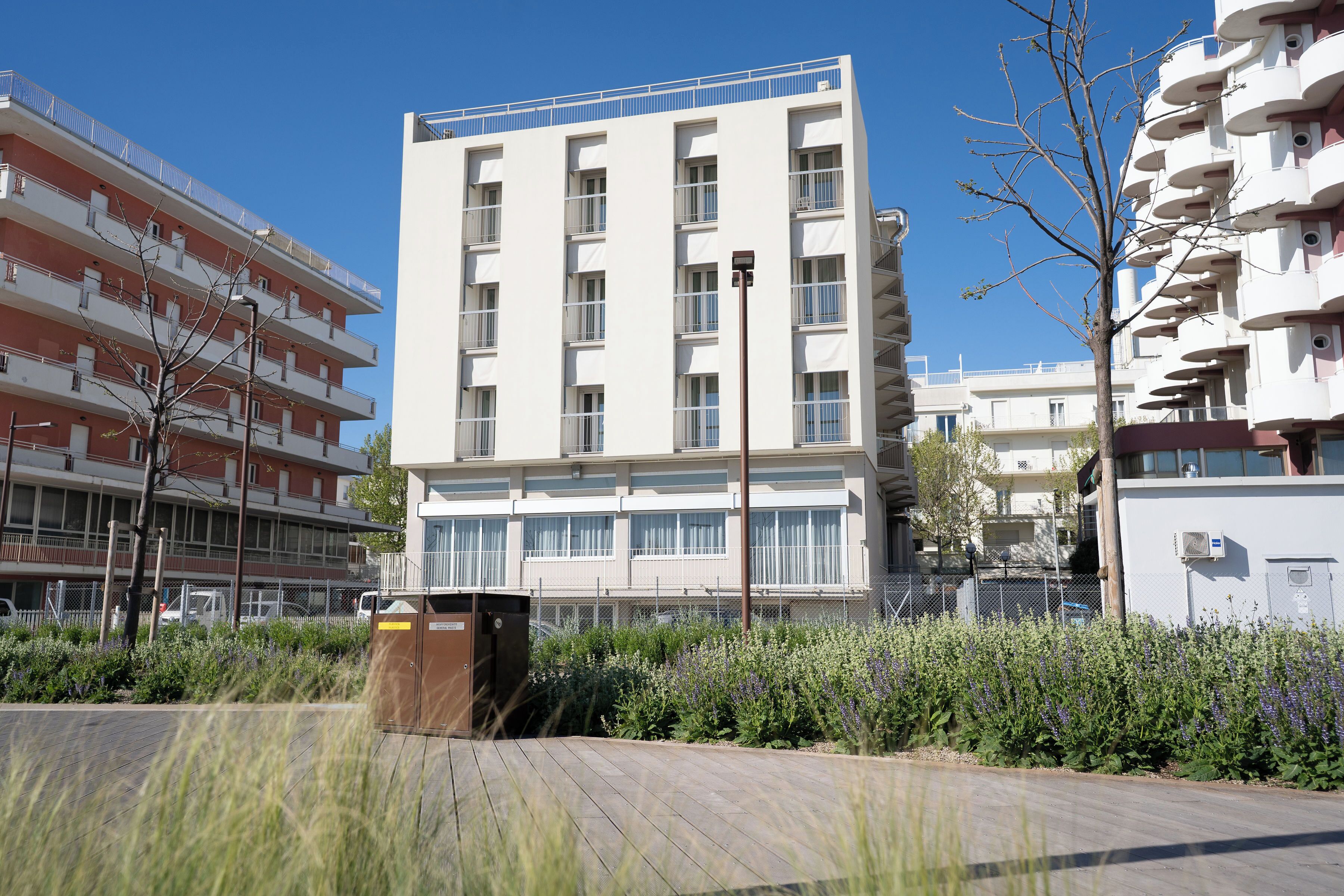 A modern multi-story building with balconies, a clear sky, and a landscaped area with plants.