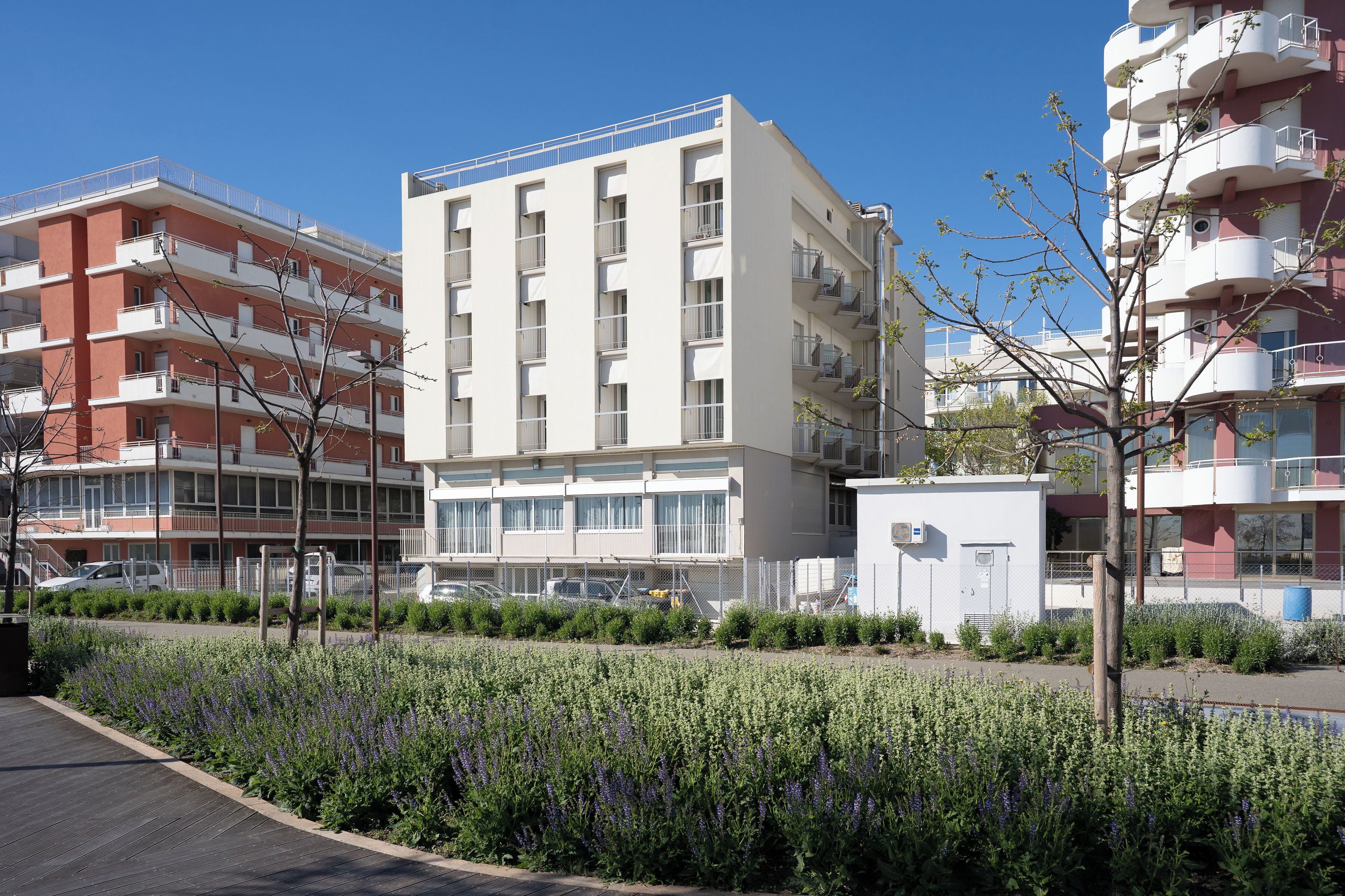 Un immeuble résidentiel à plusieurs étages, doté de balcons, d’un espace de stationnement et d’un jardin aménagé avec des fleurs violettes.