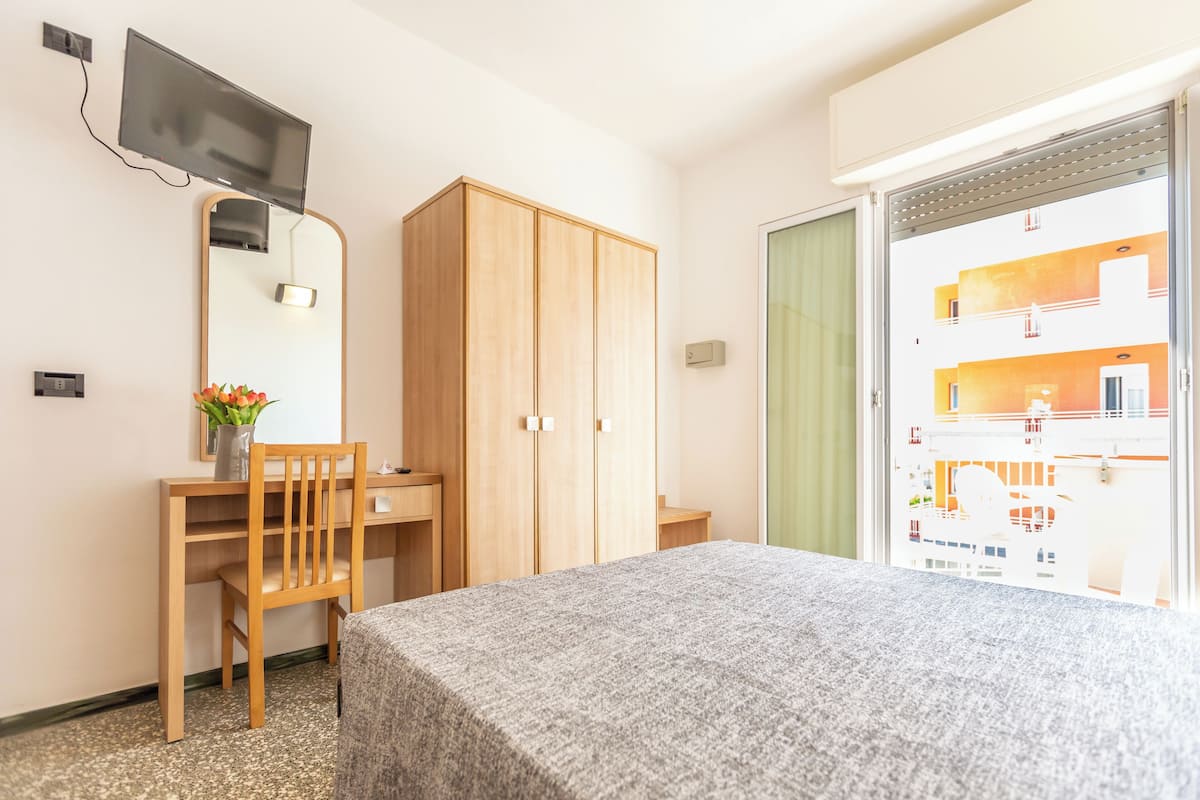 A hotel room with a bed, a desk with a chair, a wardrobe, a TV mounted on the wall, and a window with a view of the building's exterior.