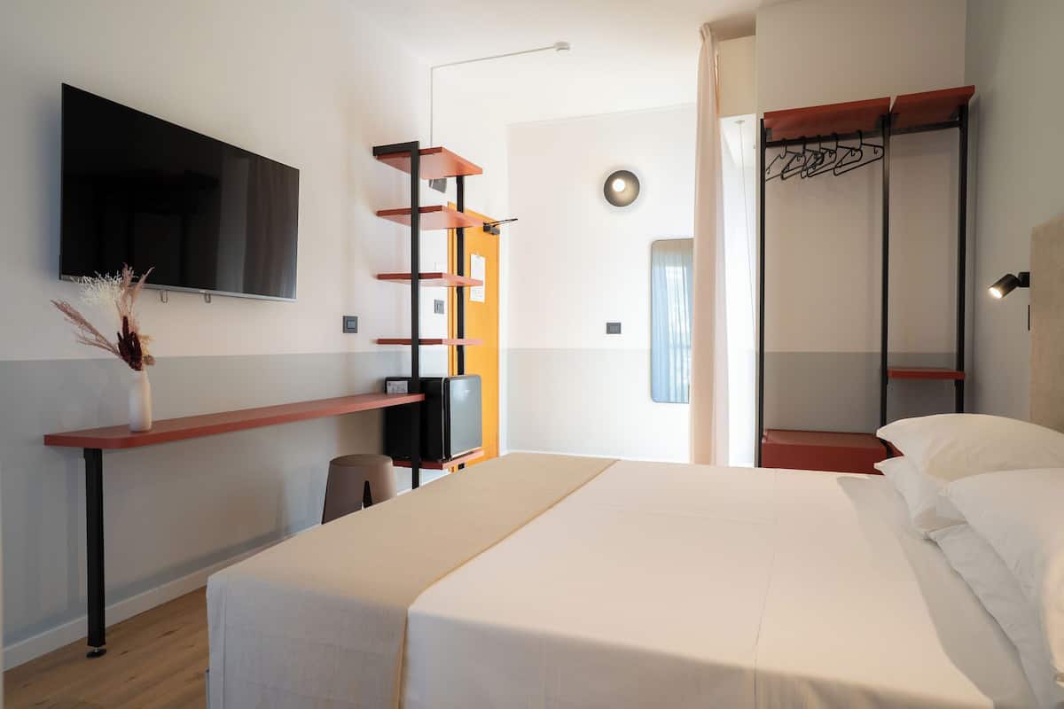 A modern hotel room with a bed, a flat-screen TV mounted on the wall, a wooden shelf, a desk with a chair, and a coat rack.