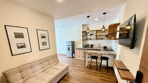 Ein modernes Wohnzimmer mit einer Couch, einem Essbereich mit Hockern, einer Küche mit Holzschränken und einem an der Wand befestigten Fernseher.