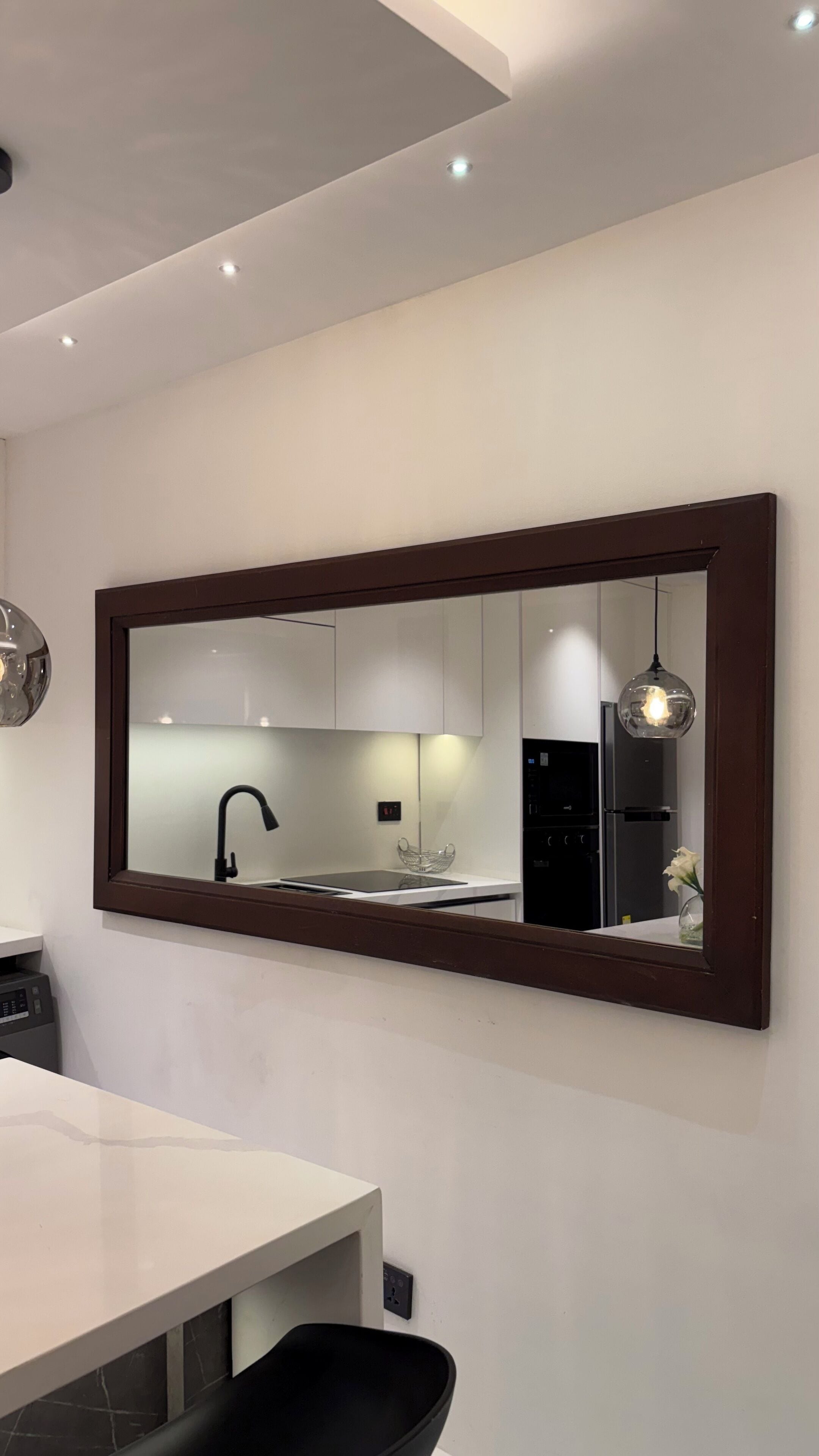 Une cuisine moderne avec un grand miroir, des meubles blancs, un réfrigérateur noir et un luminaire suspendu.