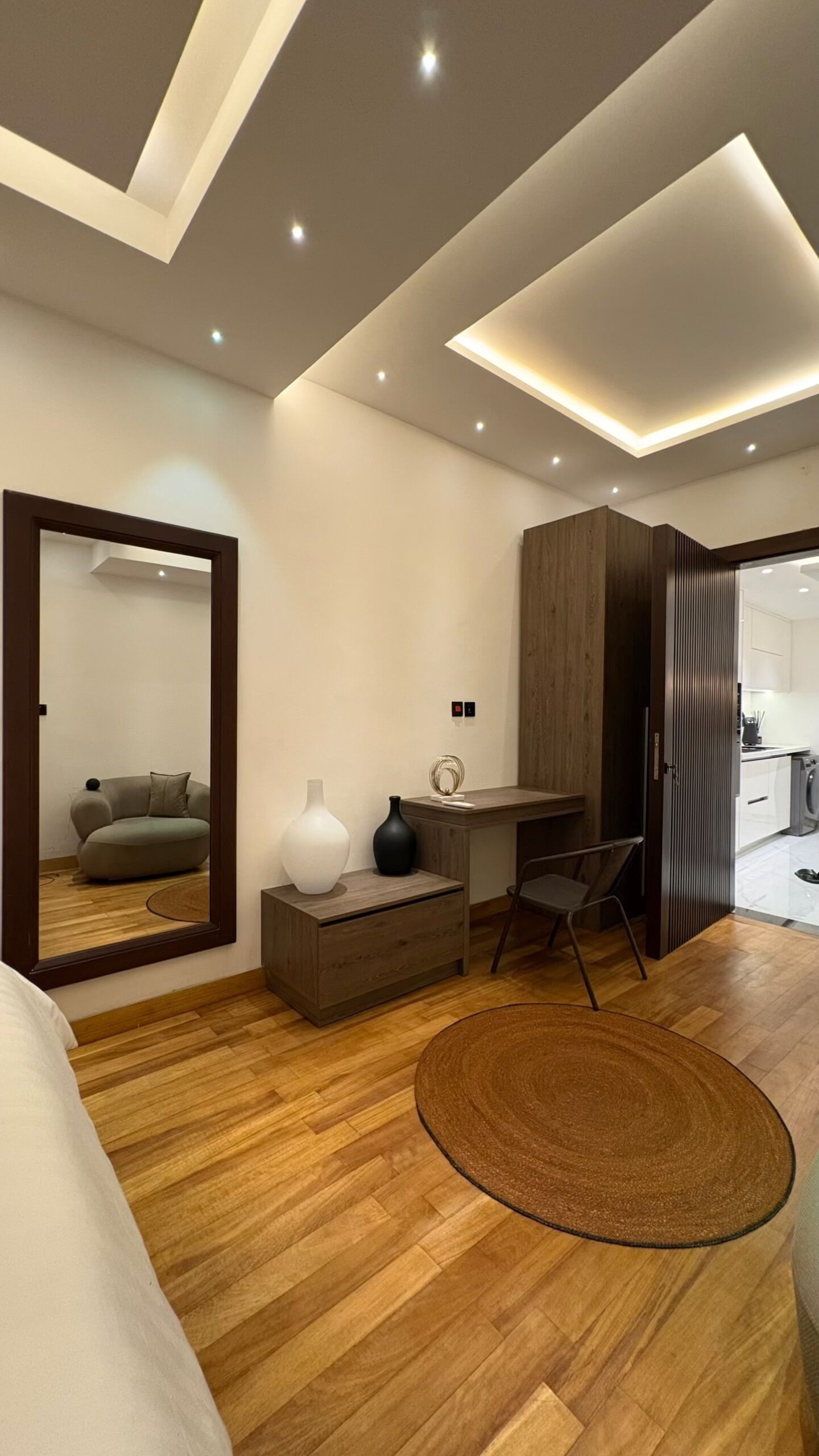 A modern bedroom with a wooden floor, a desk, a chair, a large mirror, and a round rug.