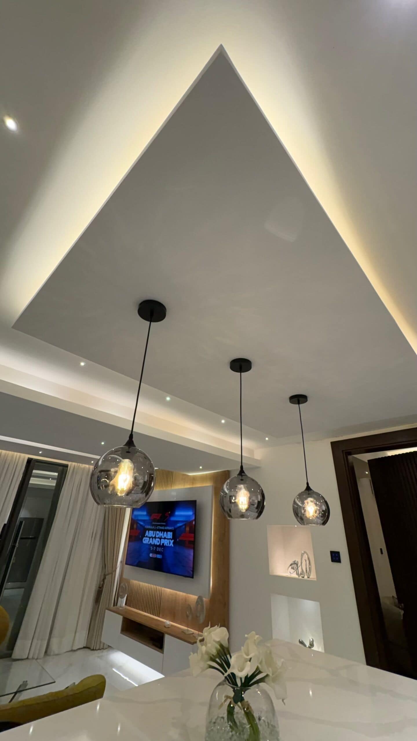 A modern living room with a high ceiling, pendant lighting, a flat-screen TV, and a marble table with a vase of flowers.