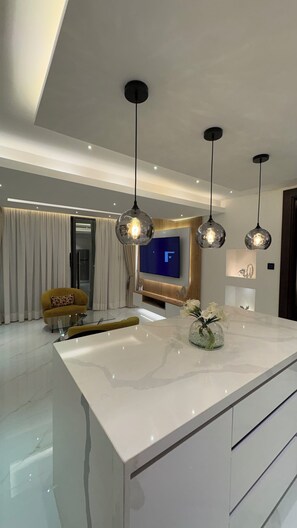 A modern living room with a large white marble island, a yellow armchair, and a flat-screen TV mounted on the wall. - Peravi Residence (Accra)
