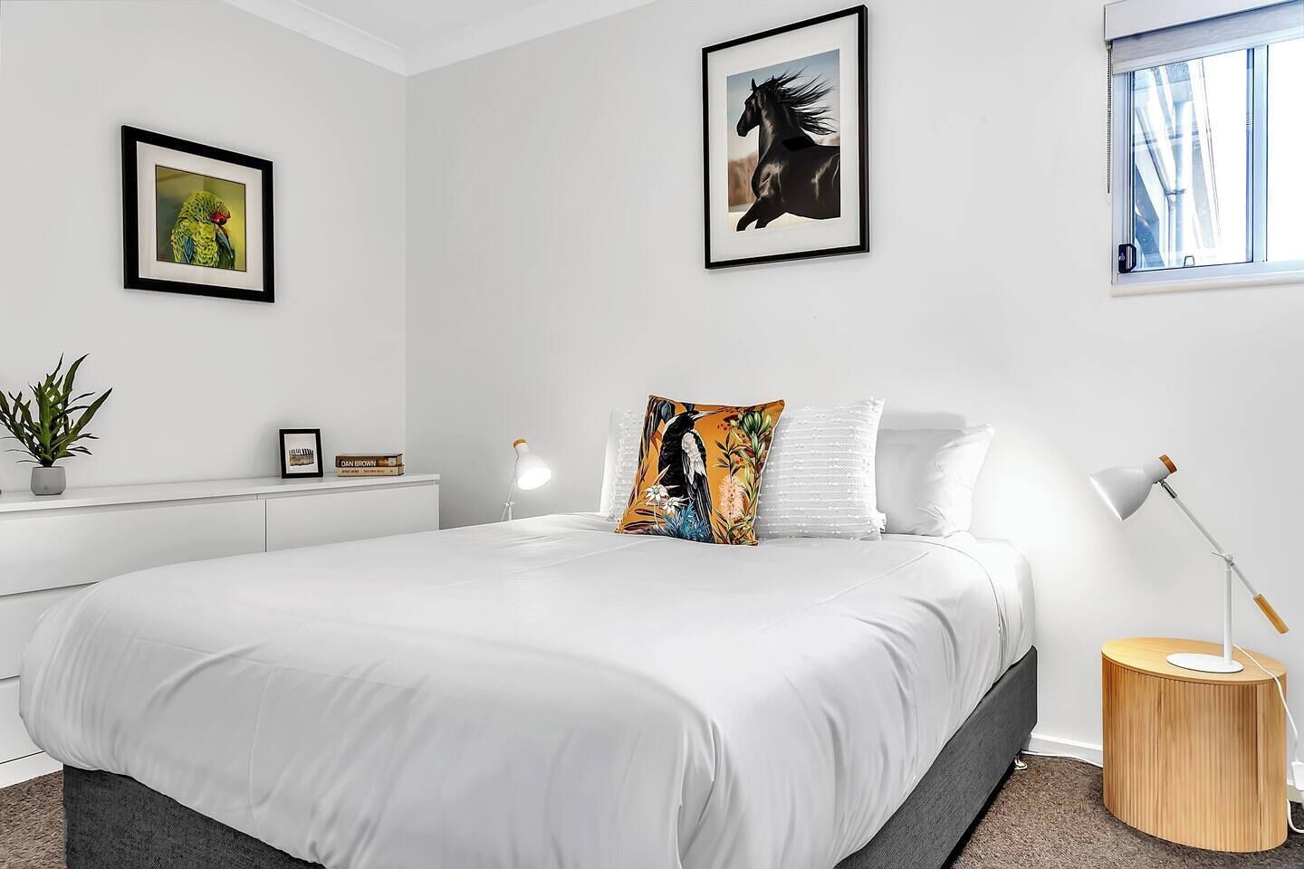 A modern bedroom with a large bed, a bedside table with a lamp, a small round wooden side table, and two framed pictures on the wall.