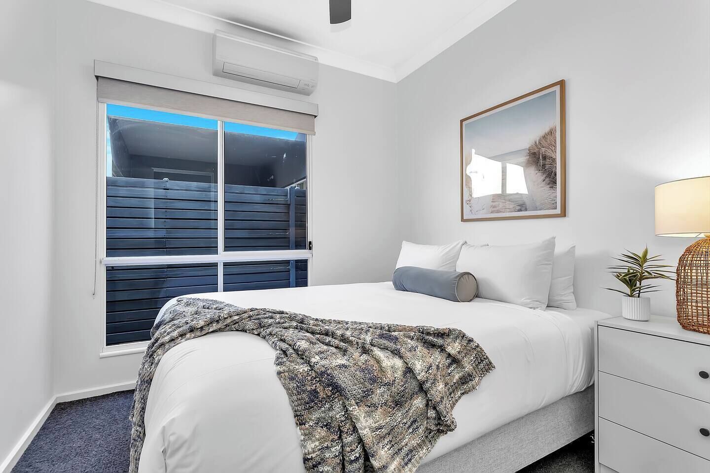 A bedroom with a large window, a bed with white bedding, a bedside table with a lamp, and a framed picture on the wall.