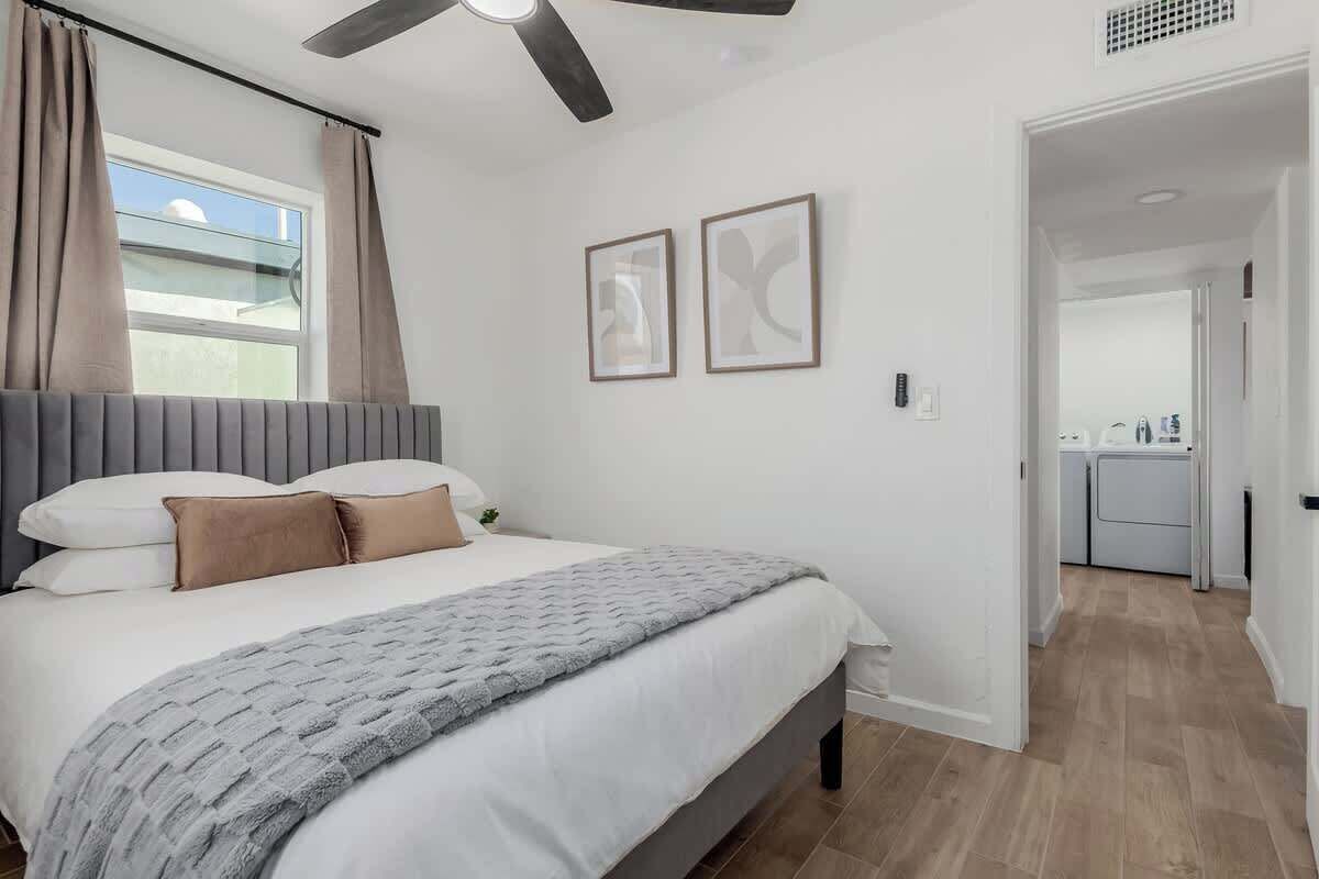 Un dormitorio con cama, ventilador de techo, ventana con cortinas y puerta que da a un baño.