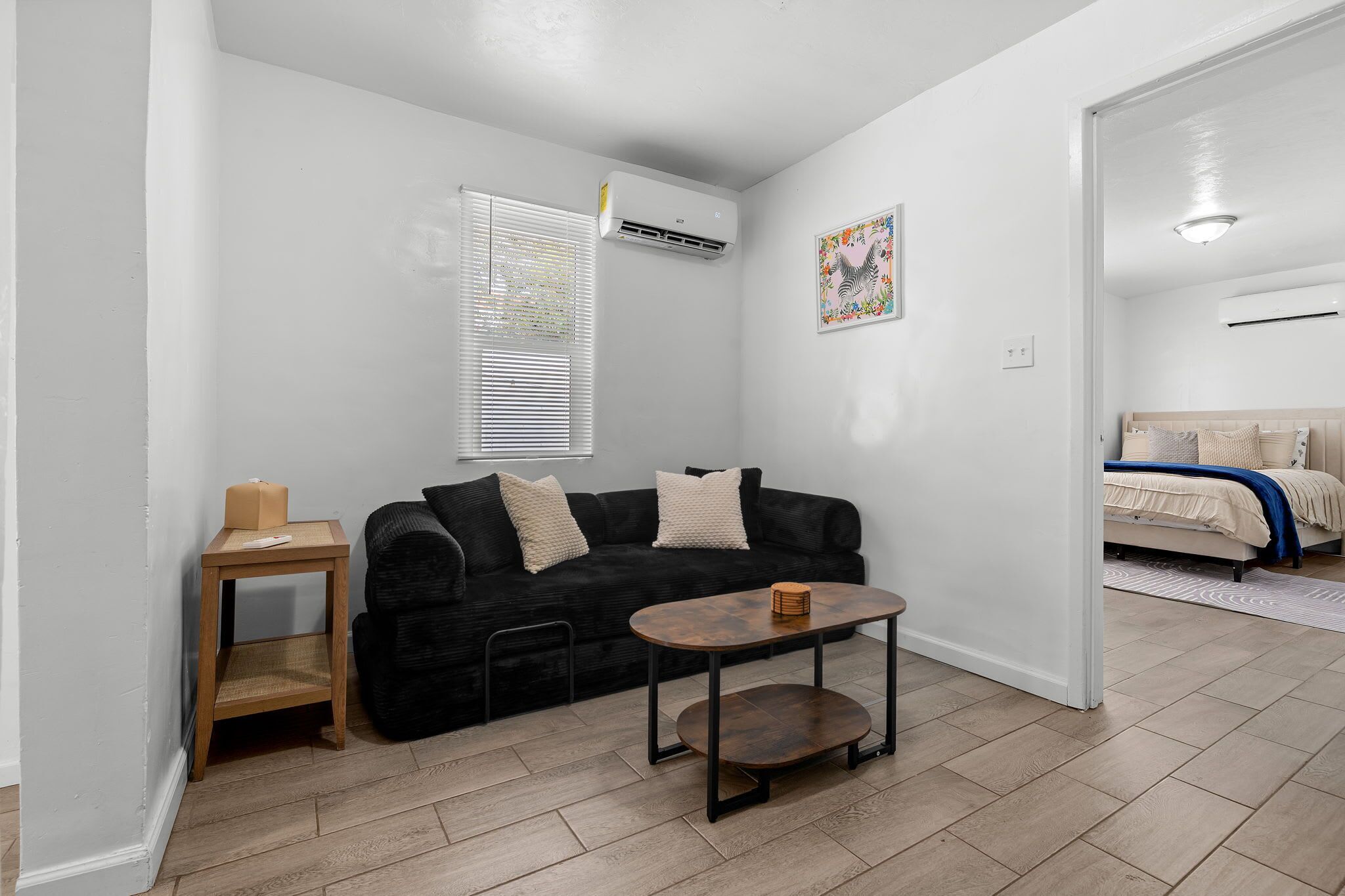 Een compacte leefruimte met een zwarte bank, een klein houten tafeltje, een enkel bed en een witte muur met één airconditioning.