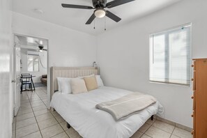 A bedroom with a bed, a ceiling fan, a window with blinds, and a door leading to another room. - Casita Rosa - Cozy - Midtown Gem (Tucson)