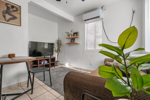 Ein modernes Wohnzimmer mit Flachbildfernseher, Esstisch, Stühlen, einer Couch, einem Blumentopf und einem Fenster mit Jalousien.